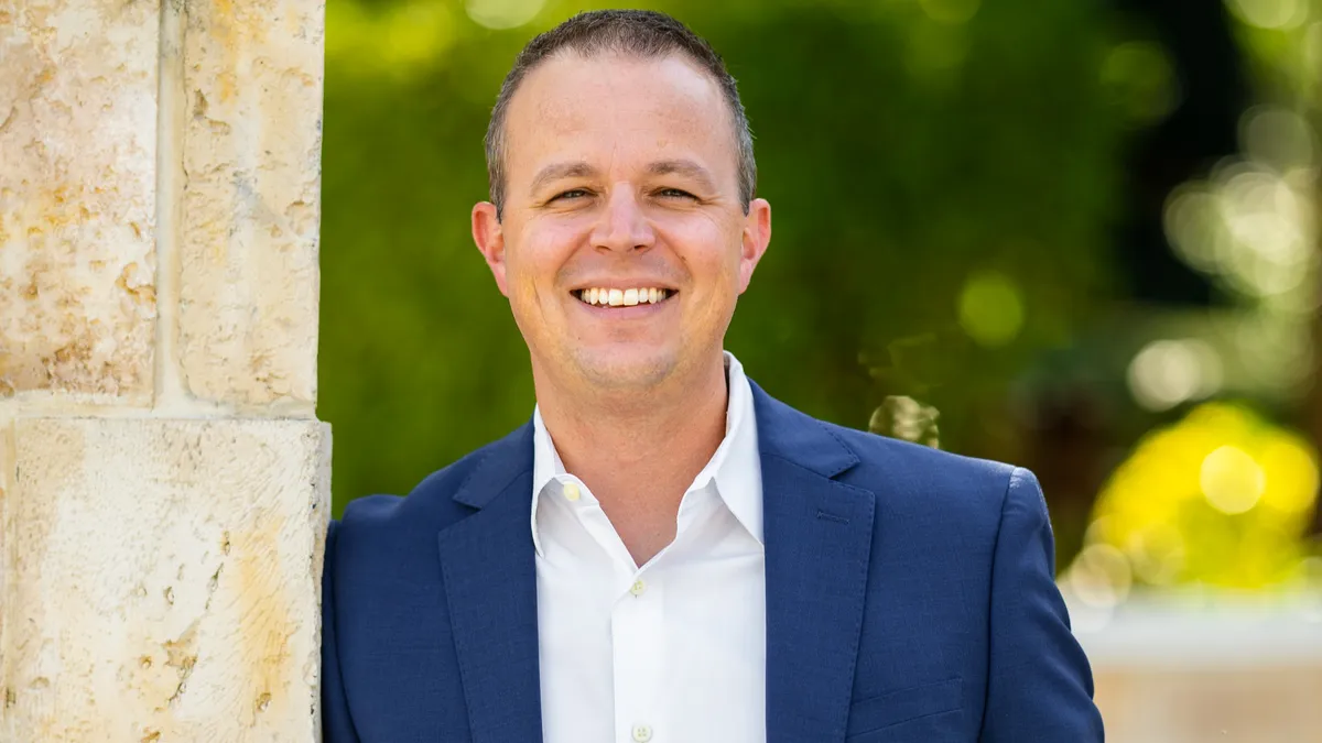 First Watch chief brand officer Matt Eisenacher wears a blue suit and leans against a beige column.