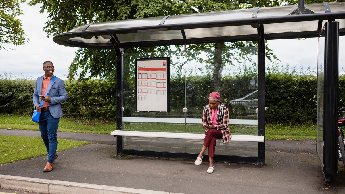 A standing man in a blue sport coat and a sitting woman wait at a bus shelter surrounded by green bushes and trees.