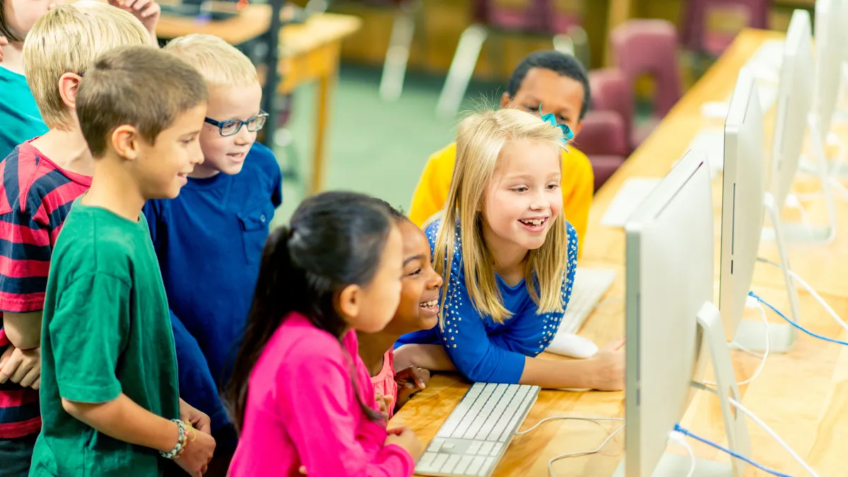 A handful of young students are in a room with desks. They are looking at one computer monitor on the desk that also has other computer monitors