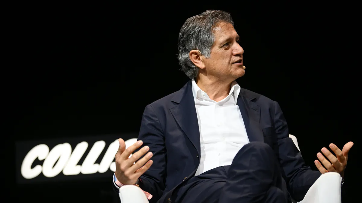 A person dressed in business attire sits in a white chair against a black background. IWhite glowing letters behind the chair read, "colli..."