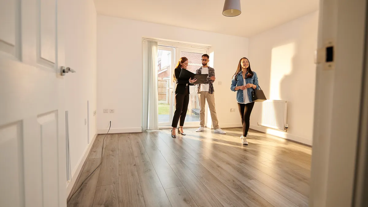 An empty apartment room with three people inside. One is showing the contents of a folder to another, while the third is walking across the room.