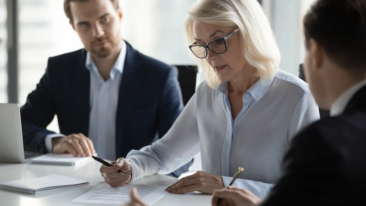 Businessmen sitting at desk headed by middle aged serious concentrated female in eyeglasses checking agreement document before signing it.