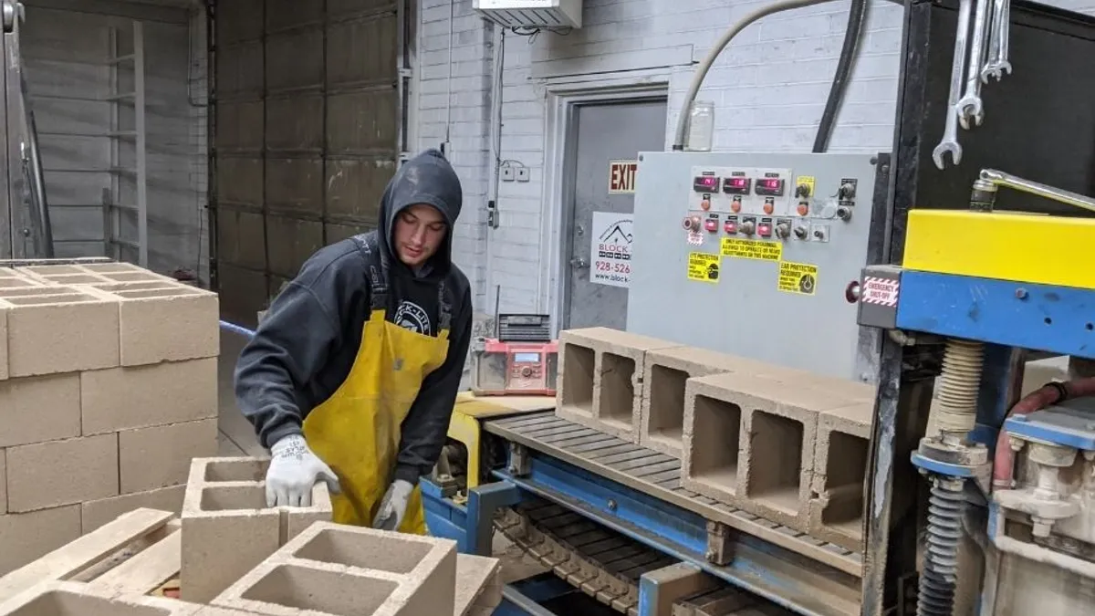 A person in a hoodie, work gloves and yellow apron picks up a block from a stack of blocks in a room with industrial equipment.