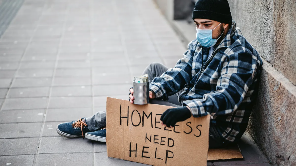 Homeless young man sitting on sidewalk and begging alone. He is wearing protective face mask.