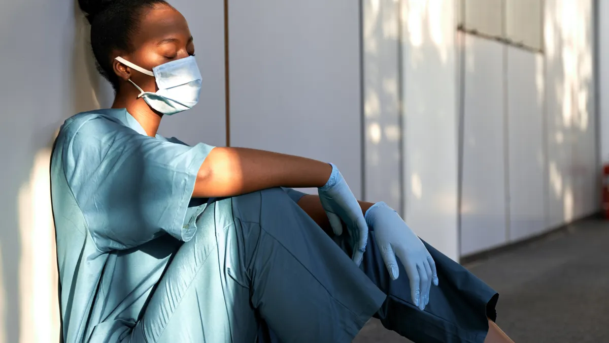 Tired exhausted female african scrub nurse wears face mask blue uniform gloves sits on hospital floor
