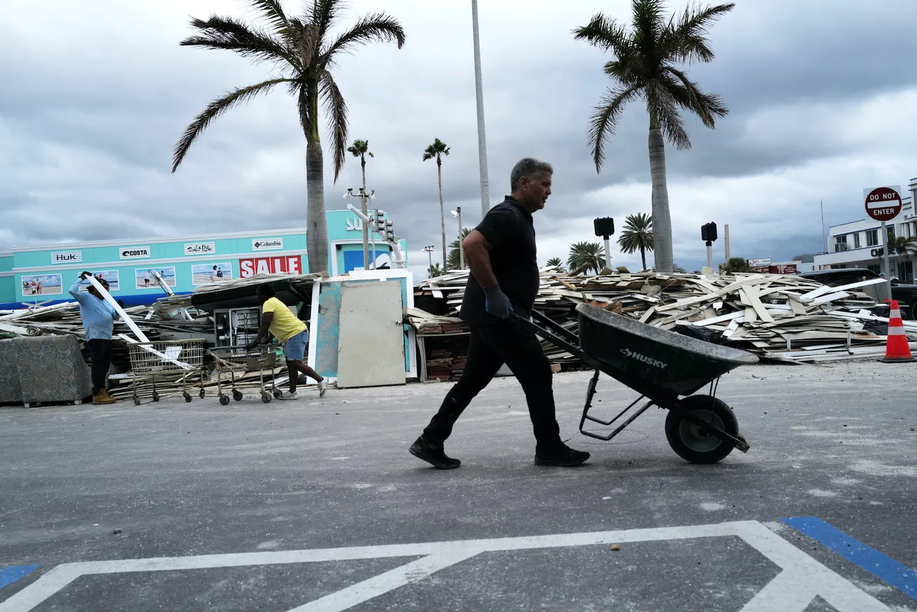 A man pushes an empty wheelbarrow with destruction from Hurricane Helene laying behind him