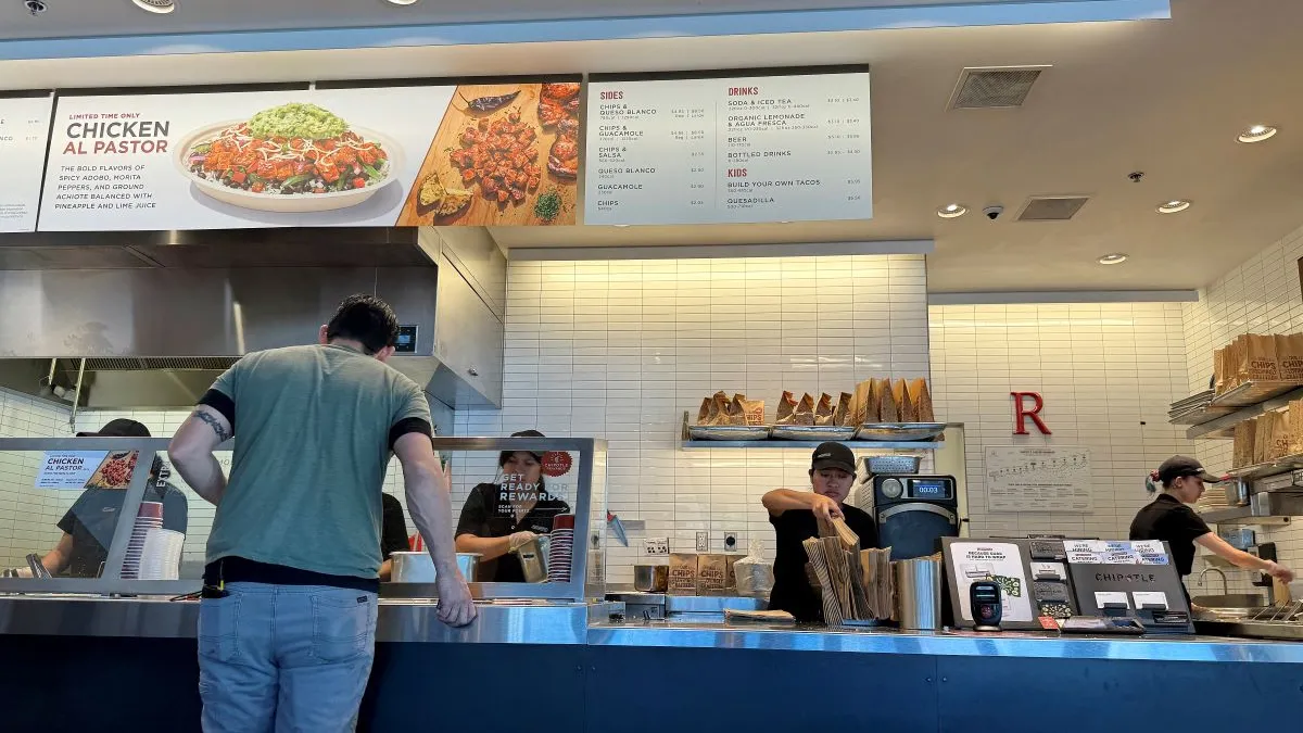 A customer in a green shirt and jeans watches workers complete a food order in a Chipotle restaurant.