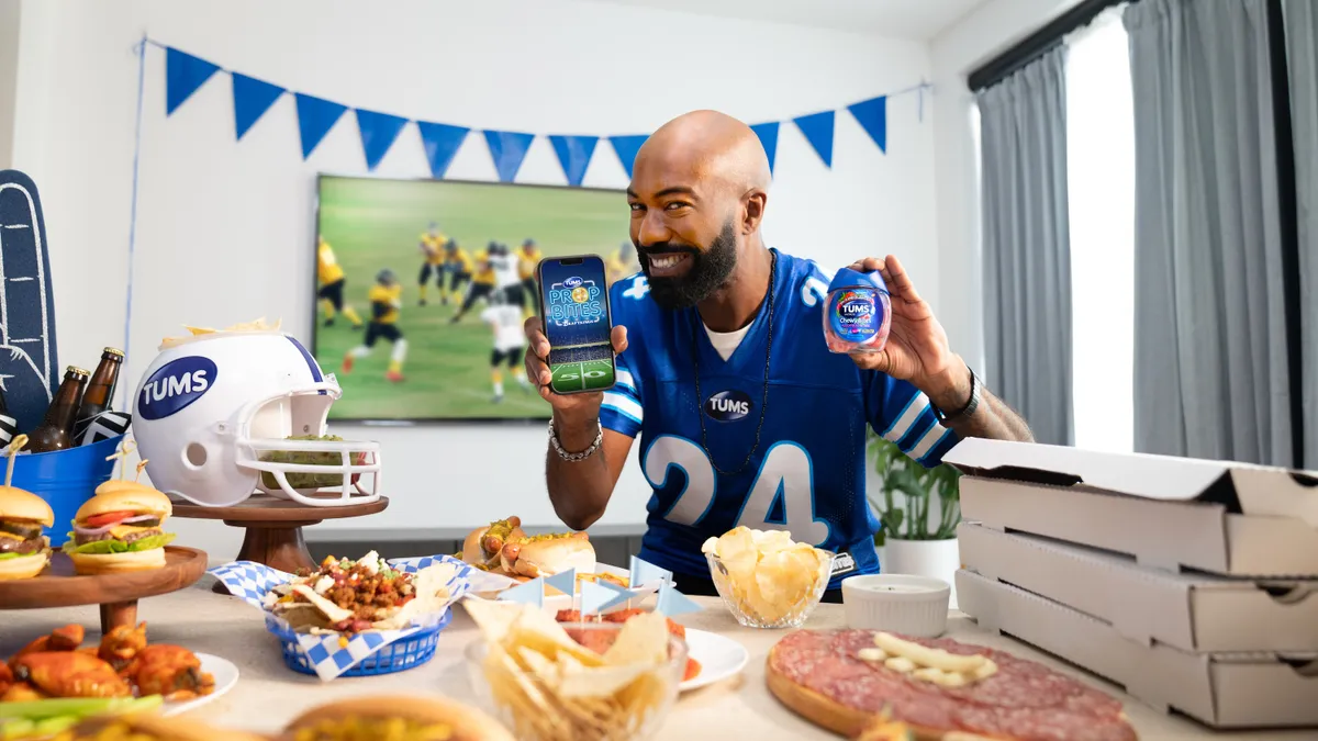 Comedian Desus Nice holds Tums and a smartphone with food on the table and football on the TV