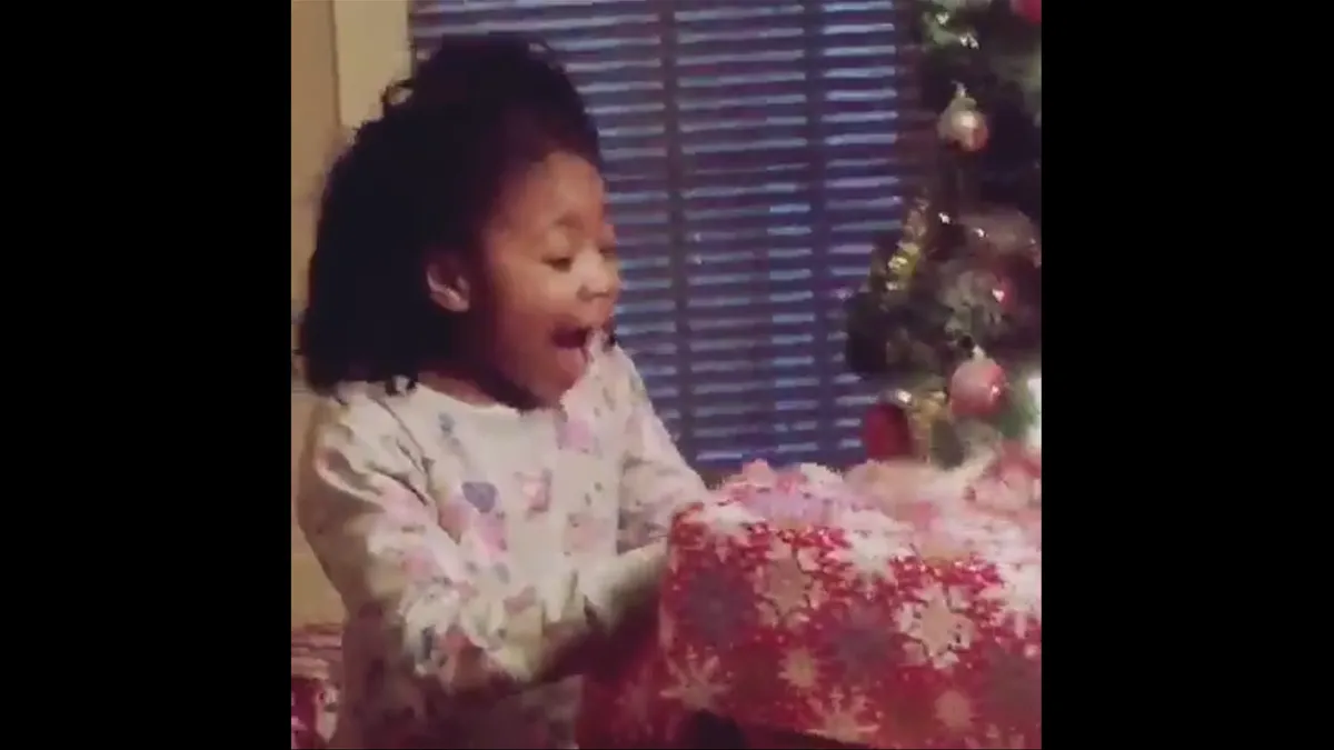 A child yells in excitement opening a present under a Christmas tree as captured on grainy home-video footage.