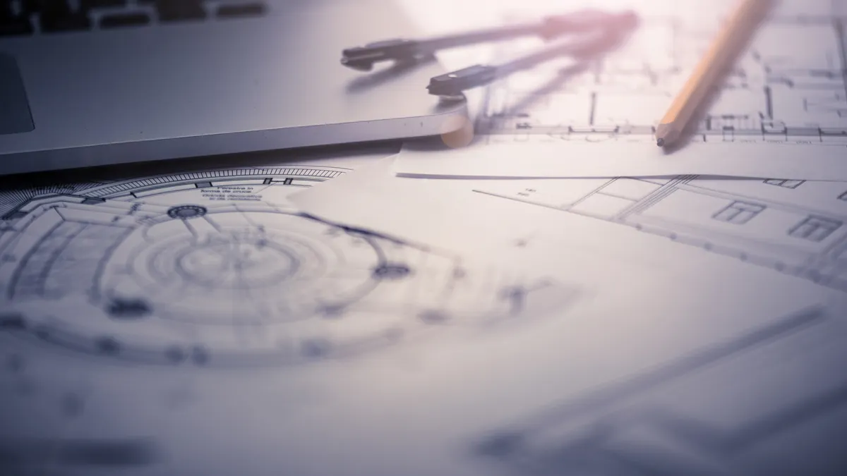 A protractor, a pencil and a building blueprint on a desk next to a laptop