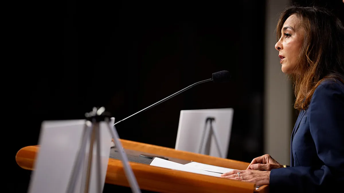 A person wearing a suit speaks into a microphone on a podium