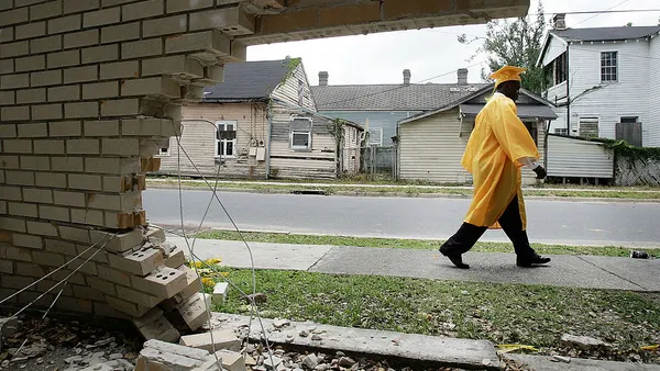 A sideview of a student walking in a graduation cap and gown on a sidewalk silhouetted by a hole in a brick wall.