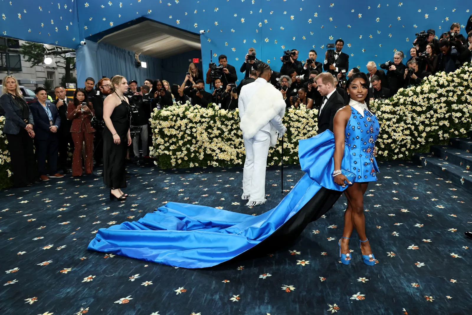 A woman in a blue minidress with a long train stands on a blue carpet.