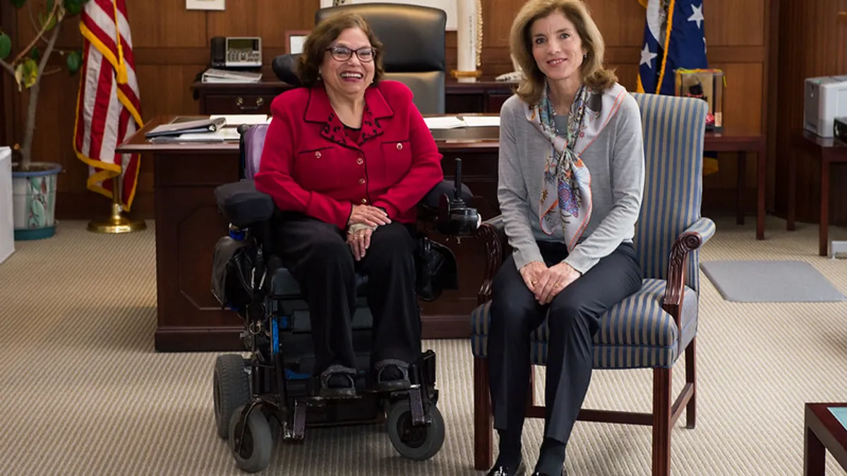 Two adults sitting in an office with wooden panelling and a desk behind them.