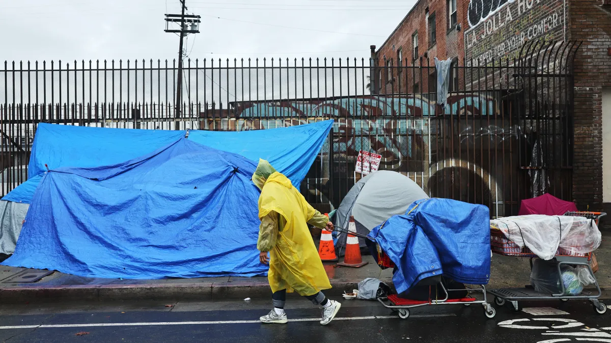 A person in a yellow rain covering pulls a cart in front of tents on a sidewalk