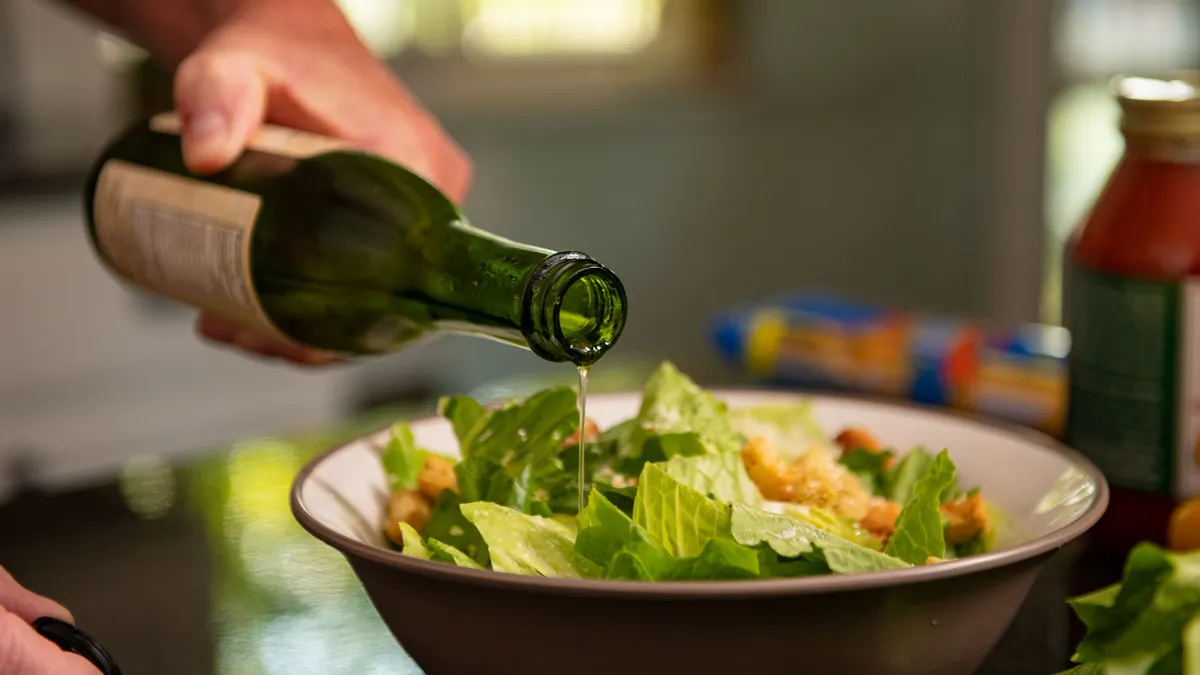 Hand pouring dressing from a glass bottle over a fresh green salad in a bowl.
