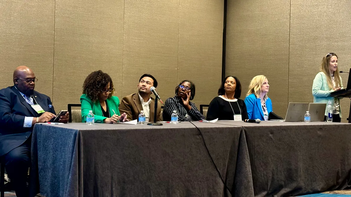 Six people are seated at a table in a conference room. Some are looking to the side at a screen that is not shown. One other person is standing near the table at a podium.