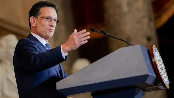 Eric Cantor, wearing glasses and a dark suit, gestures while speaking at a lectern with microphones.
