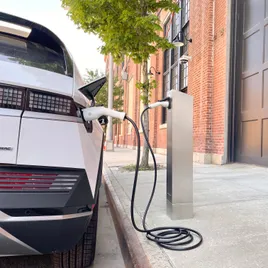 The right rear corner of a white car is parked on a street plugged in to an electric vehicle charging post.