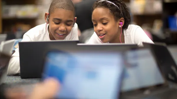 Two students with earbuds in while working together on laptops.