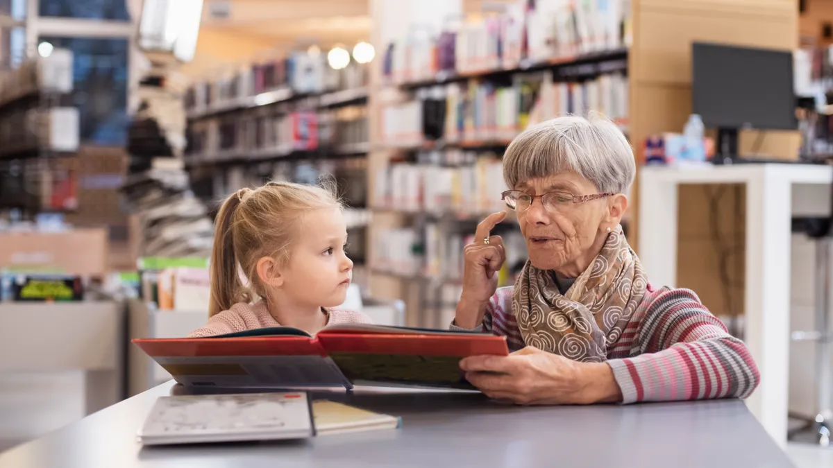 A woman from an older generation reads a book to a young girl in a library.