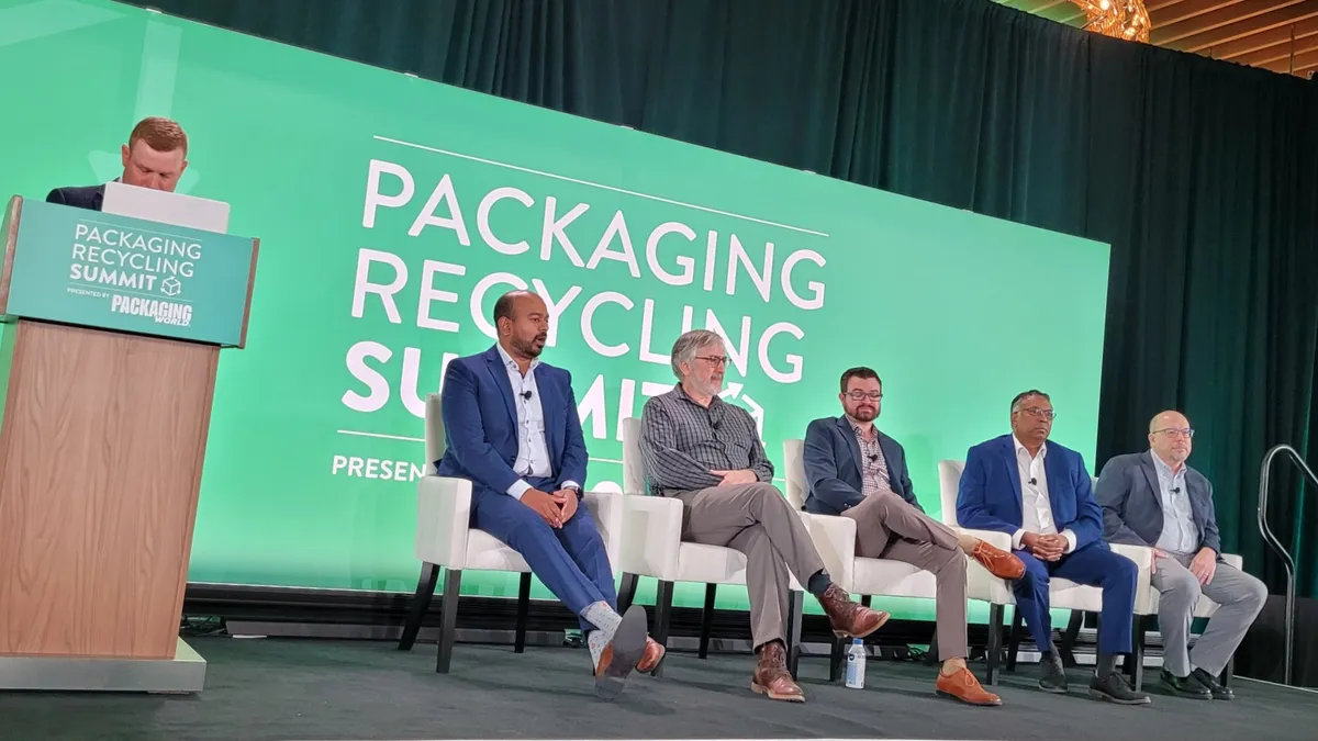 One person stands behind a podium on a stage next to five other people sitting in chairs.