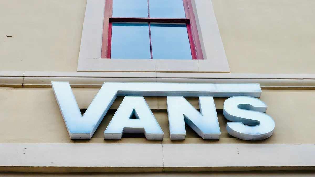 A closeup of a white "Vans" sign, below a red-trimmed window, reflecting a blue-gray sky.