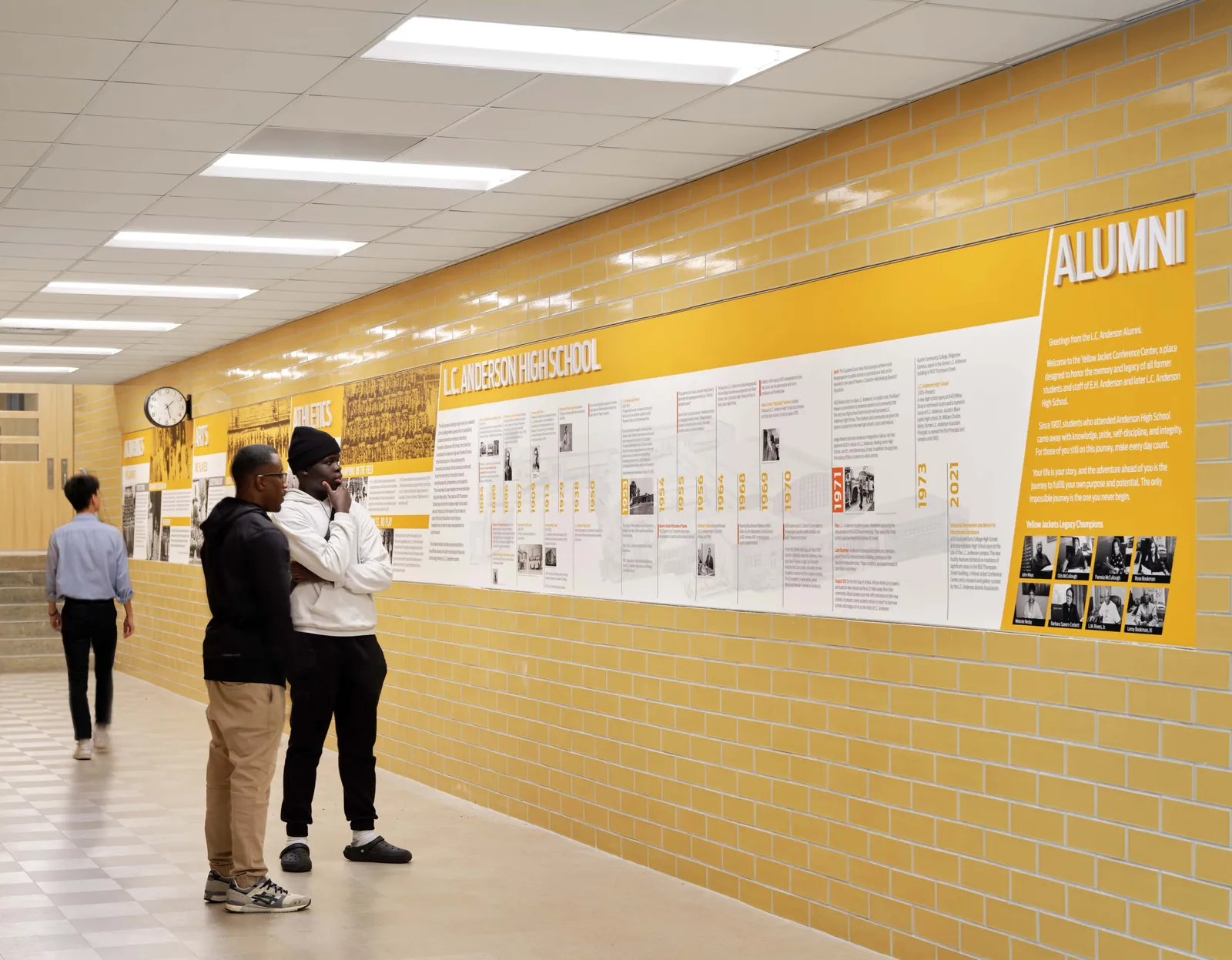 Two people are looking at a wall with writing on it inside a building hallway. Another person is walking by.