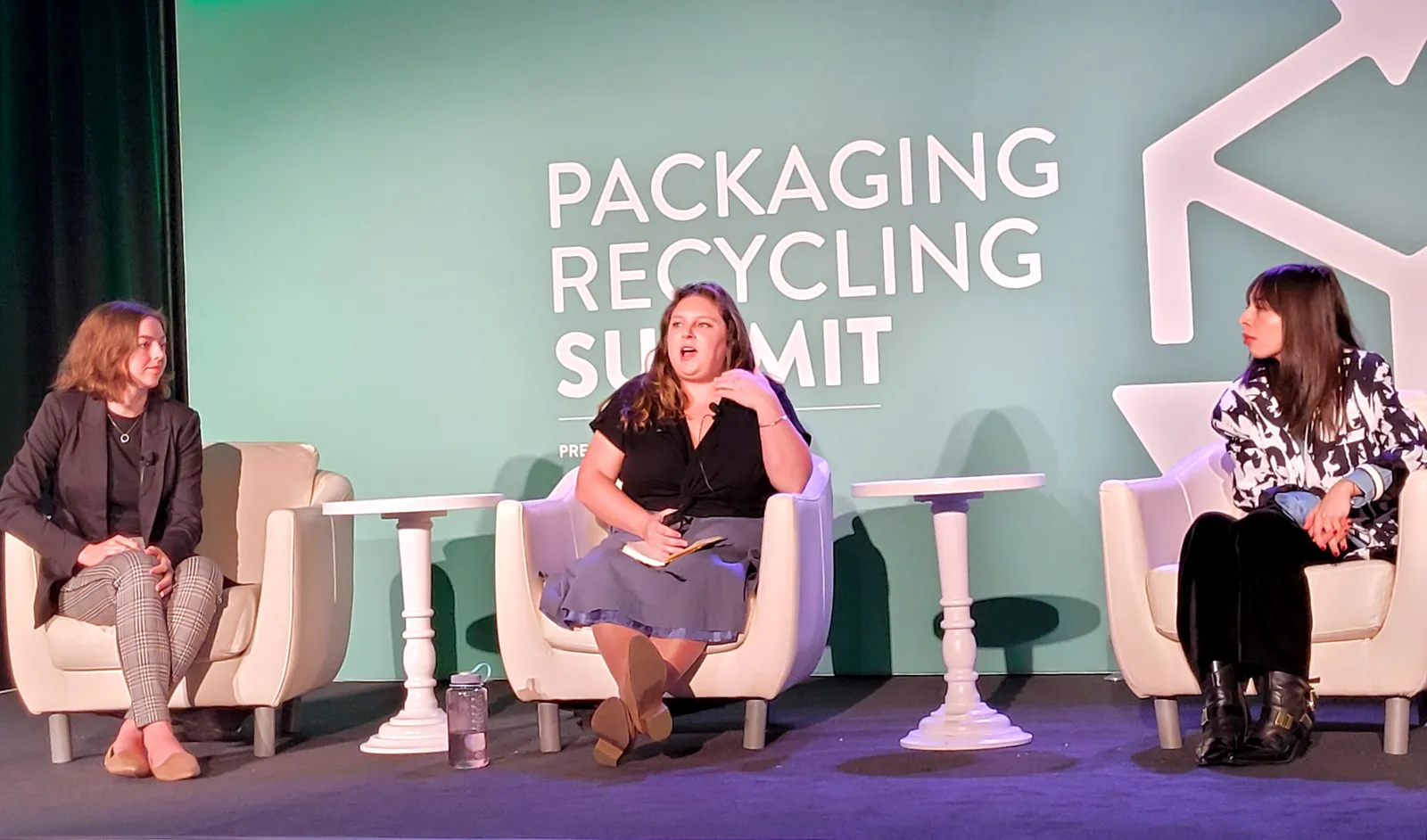 Three people sit in white chairs on a stage with a green background behind them that says "Packaging Recycling Summit."