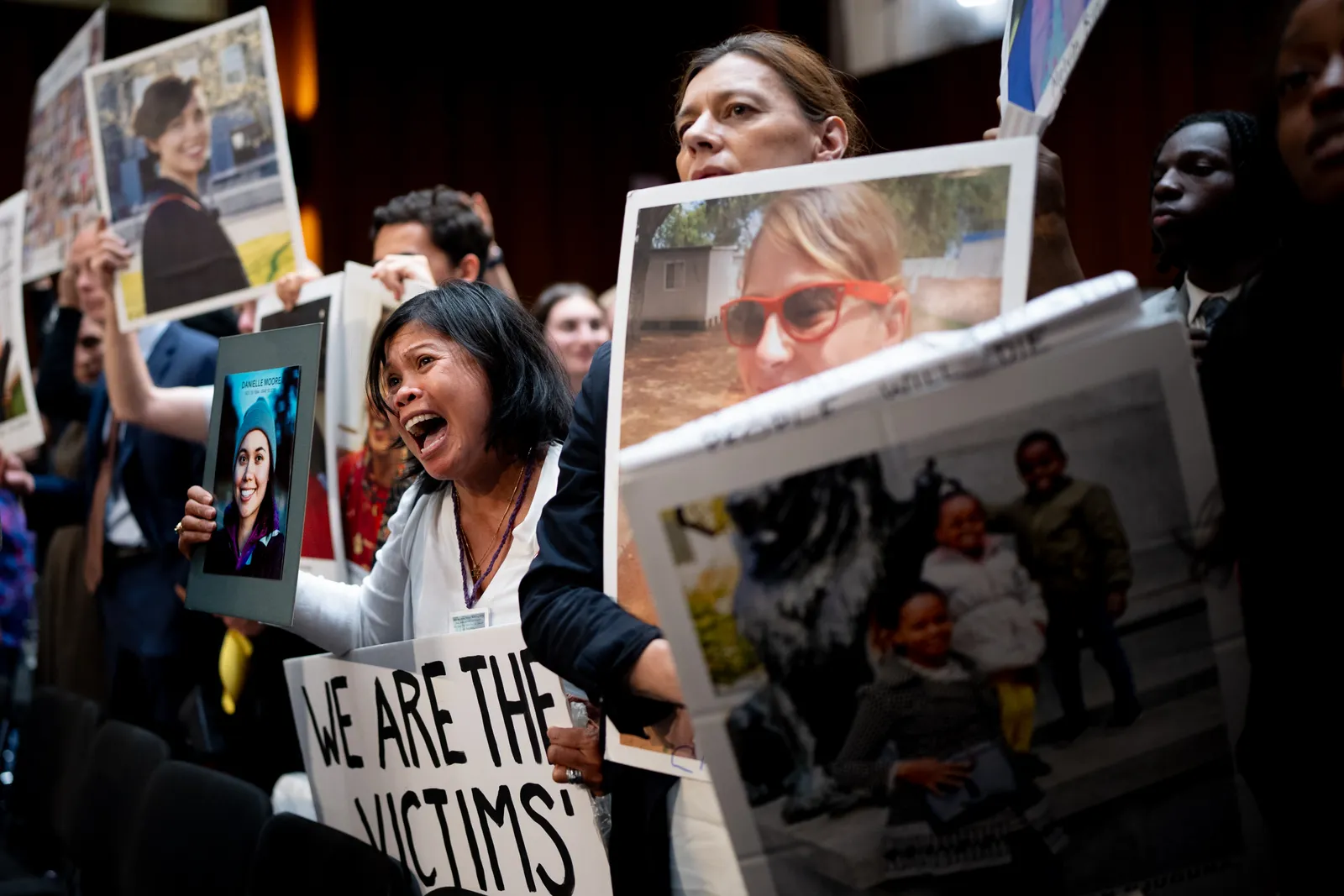 A dark-haired woman holding a picture of her daughter yelling, standing next to other people holding pictures.