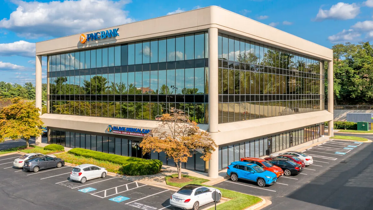 Wide exterior shot of an "All Day Medical Care Clinic" beige office building with lots of windows, surrounded by a parking lot, on a sunny day.