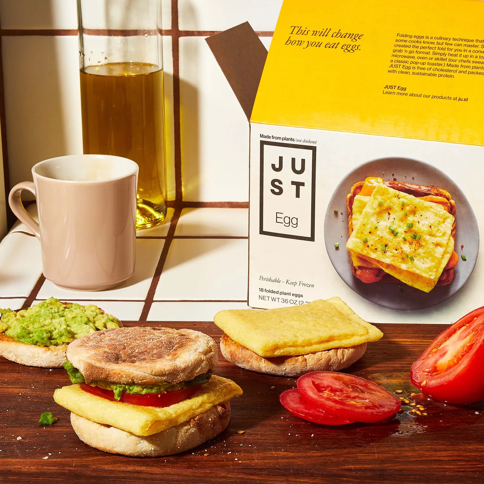 An English muffin breakfast sandwich with Just Egg folded patty, tomato and avocado and a split English muffin with a folded egg on one side and avocado on the other sit on a dark wood cutting board