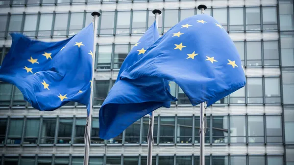 A set of three EU flags blows in the wind in front of a building.