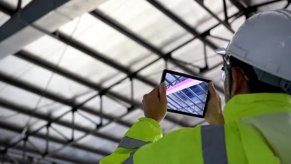 A person wearing a white hard hat and yellow-green safety vest points a tablet at an under-construction ceiling. The software in the tablet adds