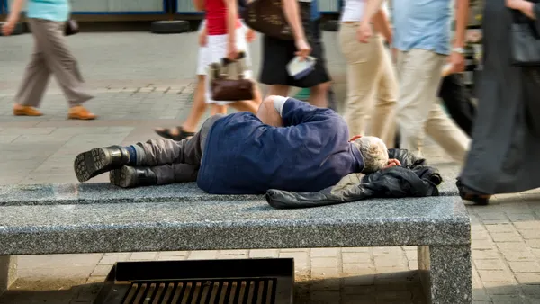 A person in a blue shirt, gray pants and boots sleeps on a bench with their back to the camera using a coat for a pillow.