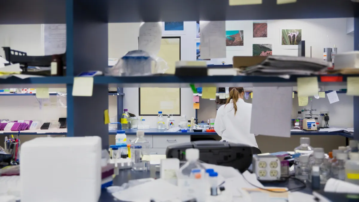 A person in a lab coat is seen through a well-lit, organized laboratory environment featuring shelves stocked with additional lab supplies and instruments. 