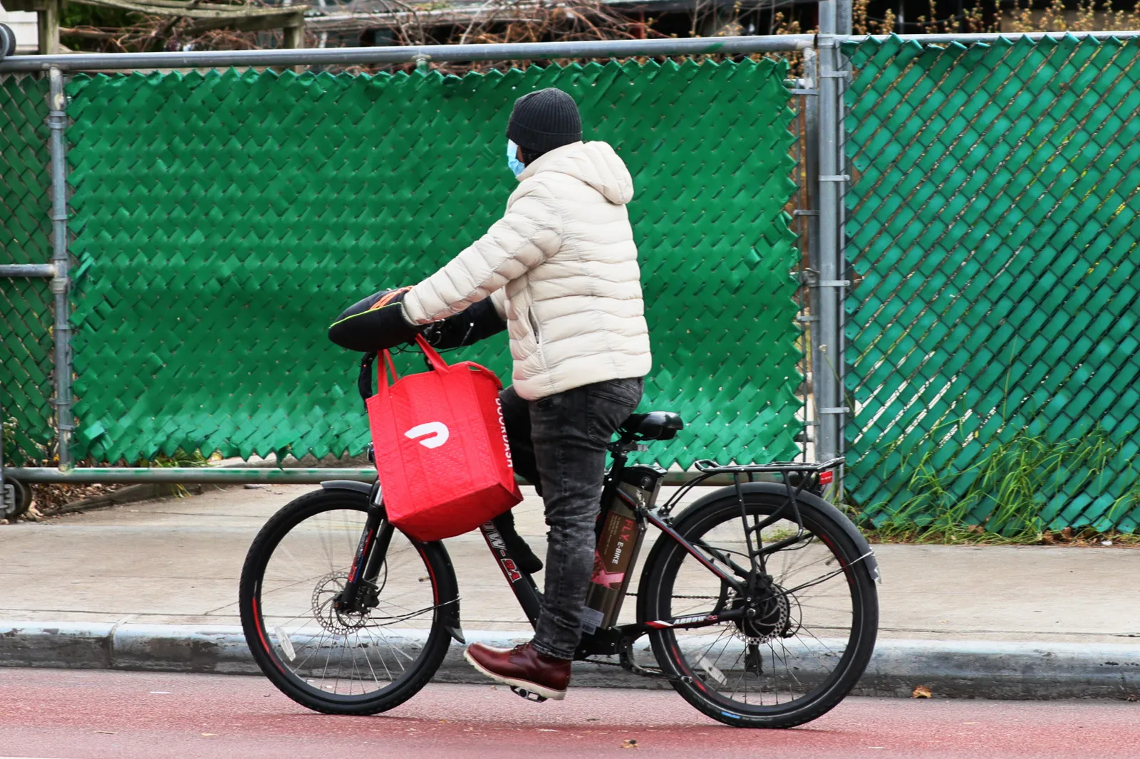 A person in a coat rides a bicycle holding a red DoorDash bag.