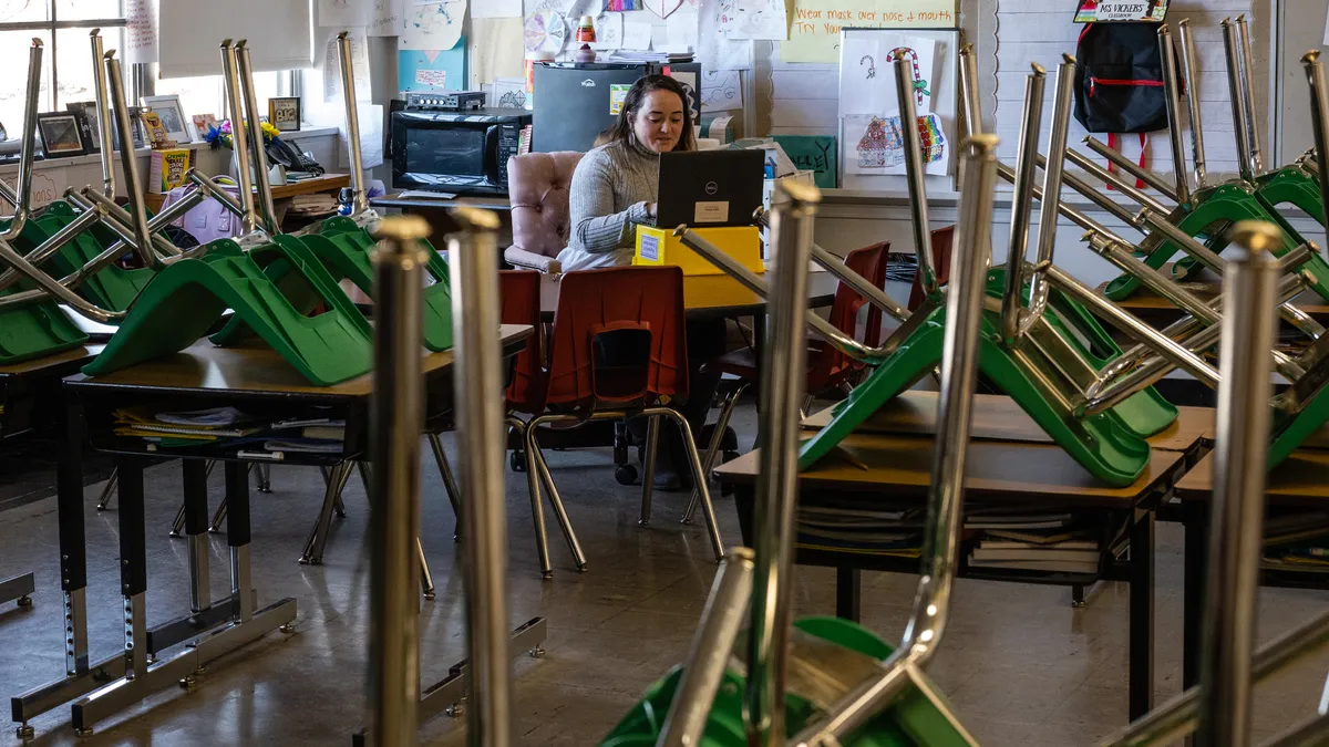 An adult sits at a desk in a classroom with a laptop open. In front of the adult are desks with green chairs sitting upside down on the desks.