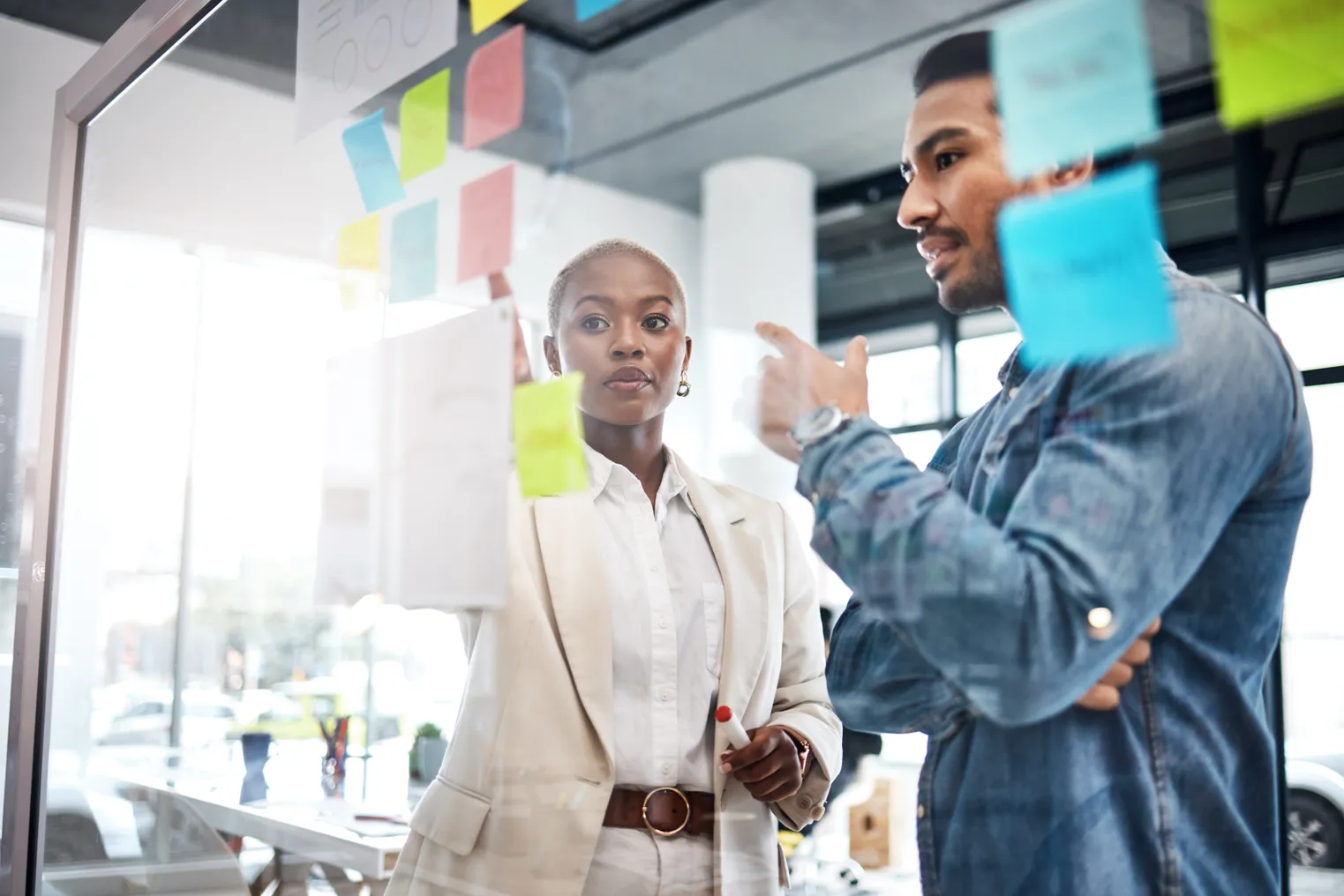 Two people in business attire stand front of a glass wall in an office with sticky notes on it, having a conversation.