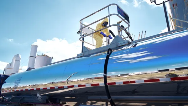 A person wearing a hazmat suit stands on top of and dips a probe into a large metal tank.