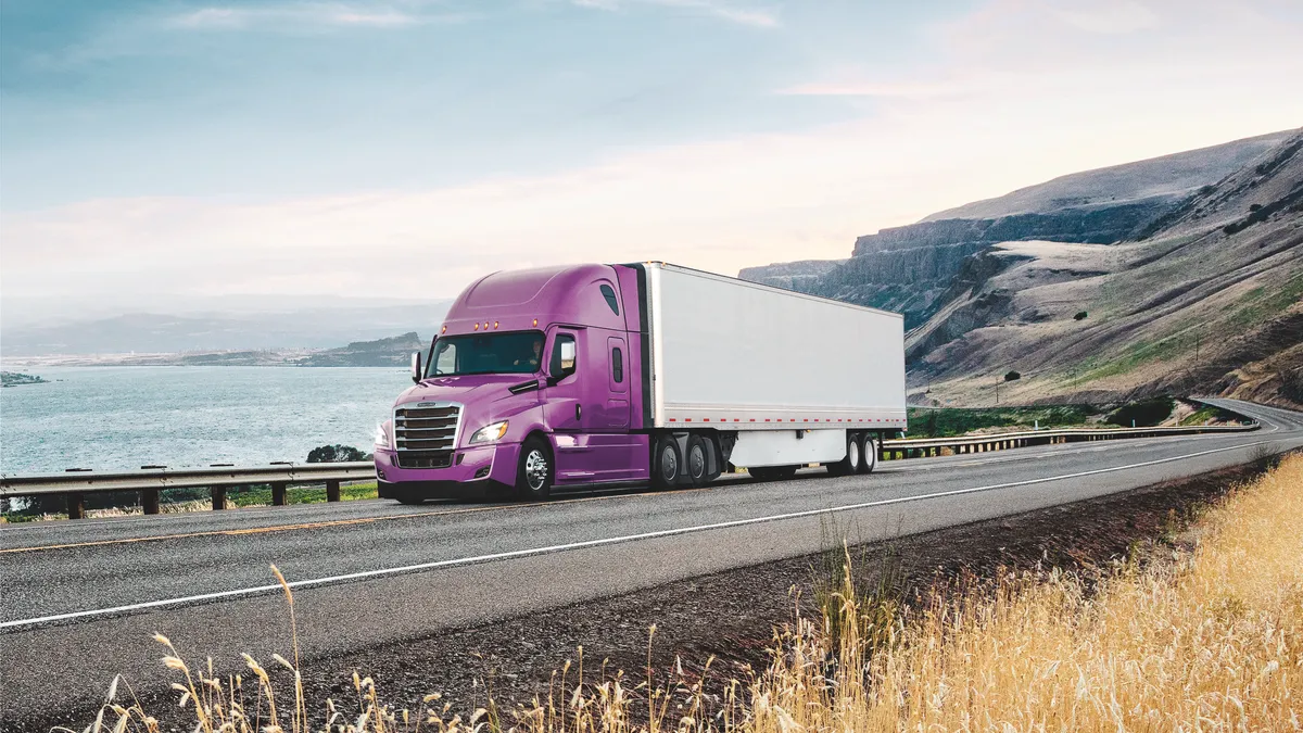 A purple Cascadia tractor pulls a white trailer next to a field and water.