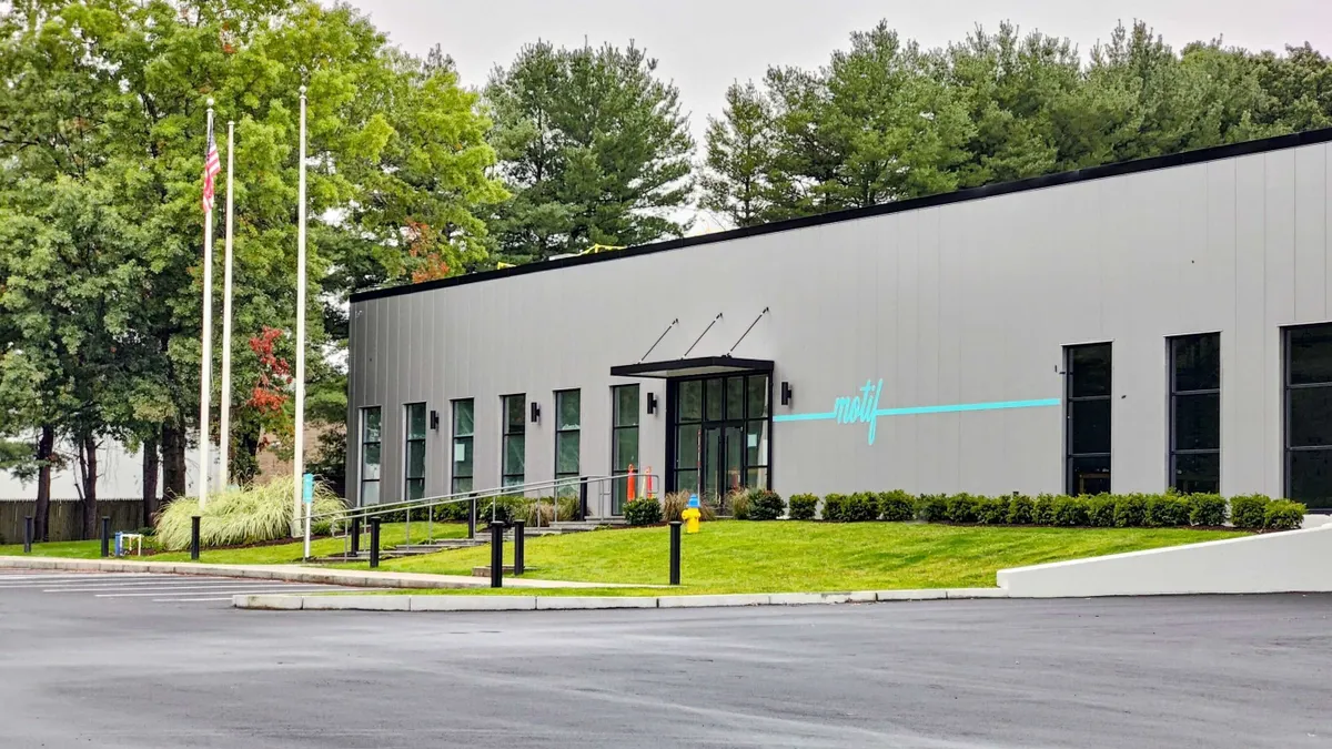 The entrance of a grey, rectangular building with a cyan blue Motif logo on it. There's an empty parking lot and green lawn in front, trees behind it, and several dark windows.