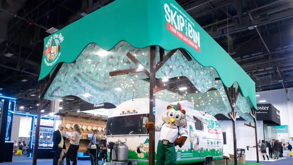 A person in a Woodsy Owl costume stands in front of an Airstream trailer at a convention center booth