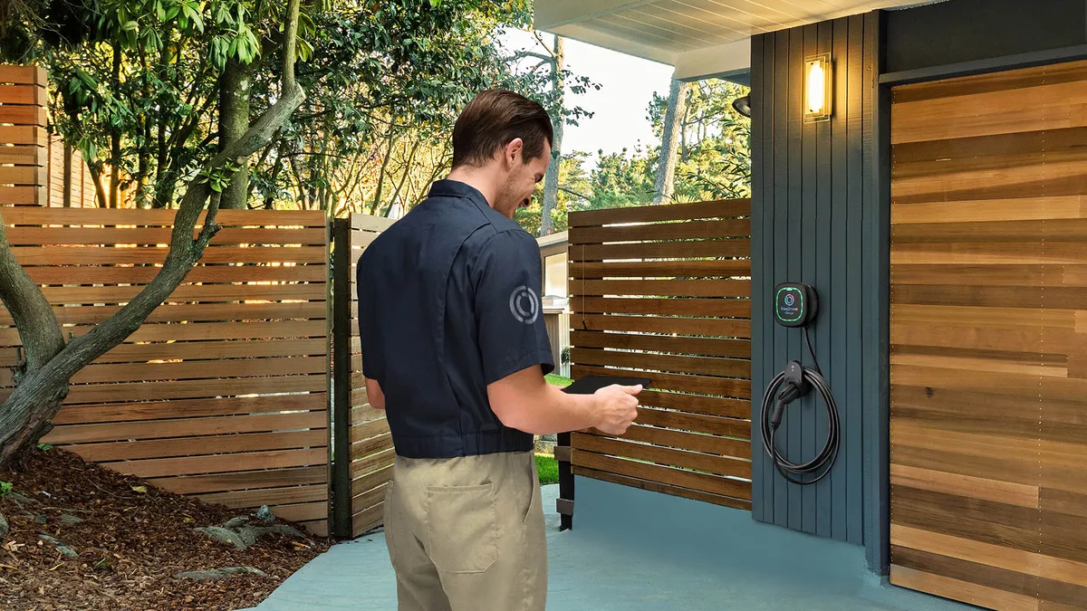 A service person looks at a tablet computer while standing in front of a garage with a wall-mounted electric vehicle charger in the background.