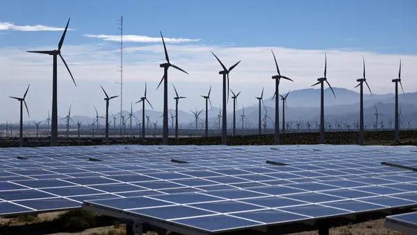 Solar panels stand in front of a row of wind turbines.