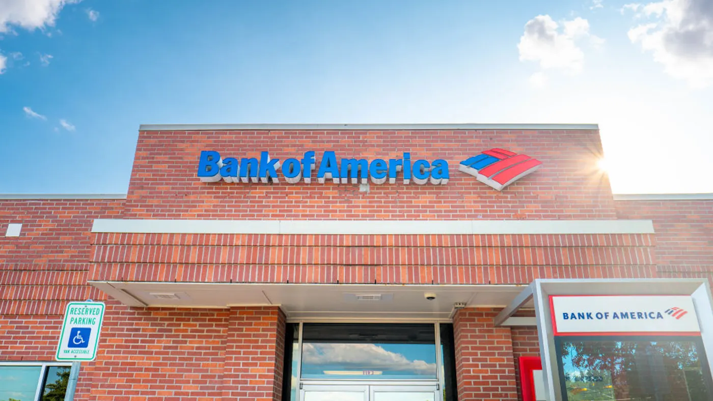 A bank branch building features the "Bank of America" name and logo in front of sunny skies.