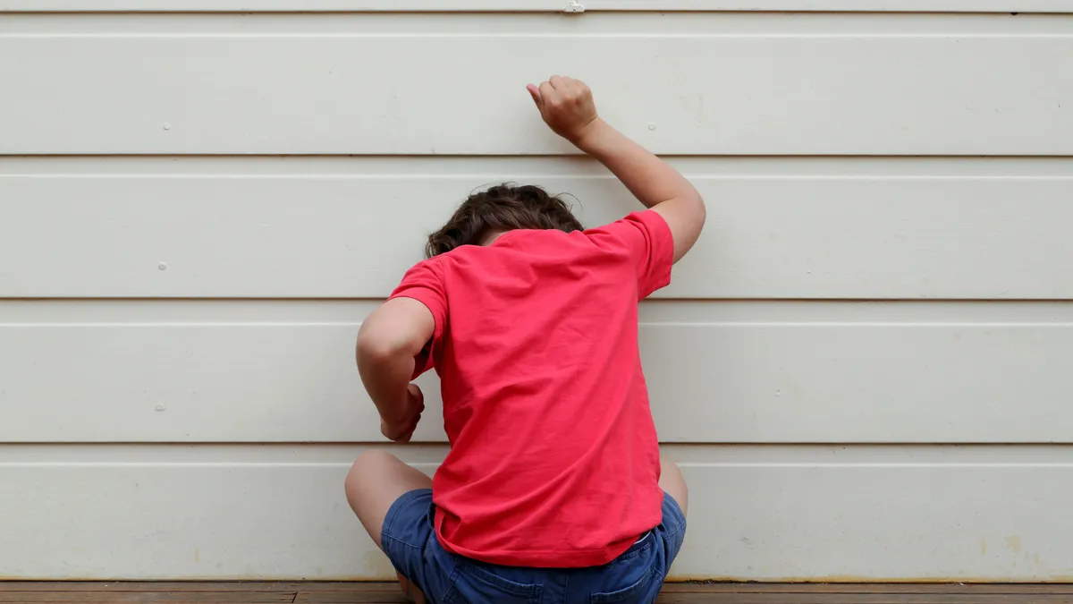 A child in a red shirt sits with face against wall and back toward camera. One hand is up with clenched fist.