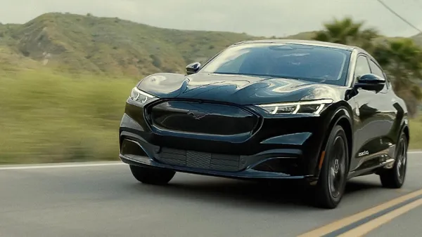 A 2023 Ford Mustang Mach E electric SUV driving on a two-lane road with green hills in the background.