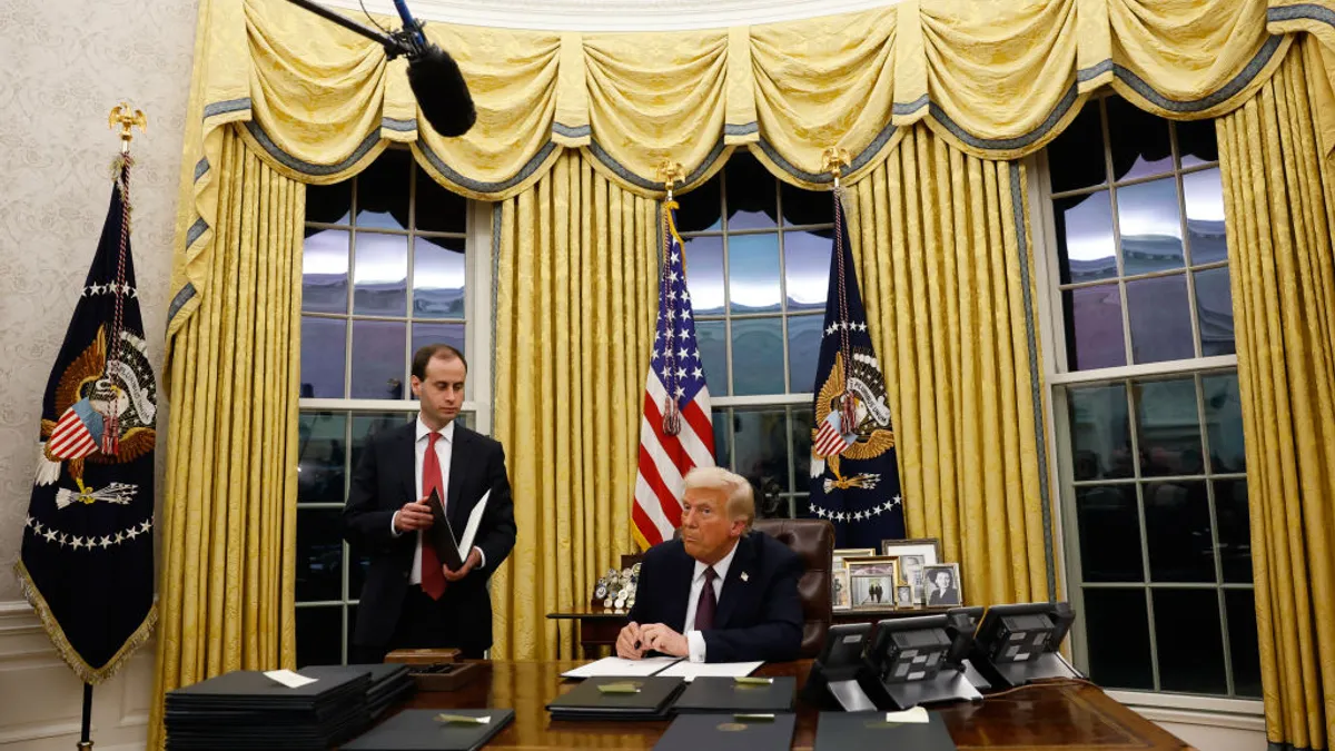 Donald Trump sits at a desk in the Oval Office. A person is standing next to the desk. There are several folders on the desk.