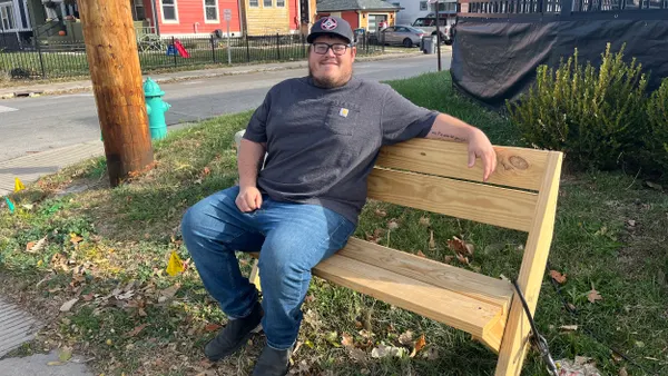 A person sits on a wooden bench on the grass with houses behind.